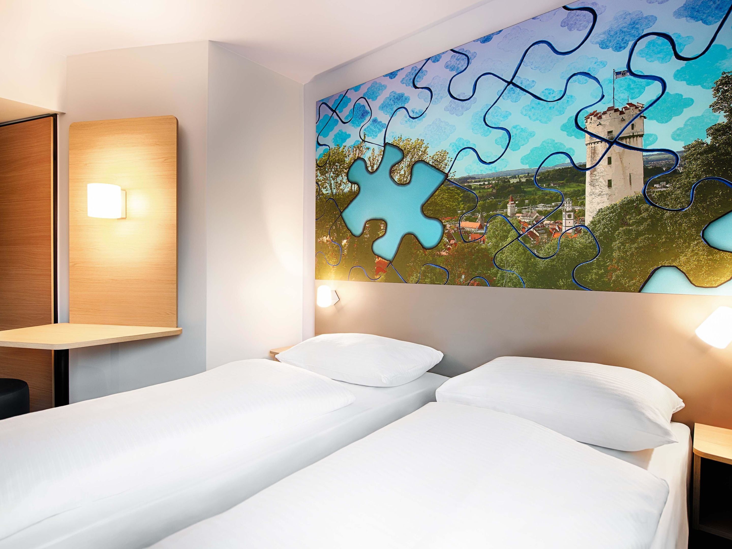 A hotel room with two single beds, a wooden headboard, a wall-mounted light, and a large puzzle-style artwork depicting a castle and landscape.