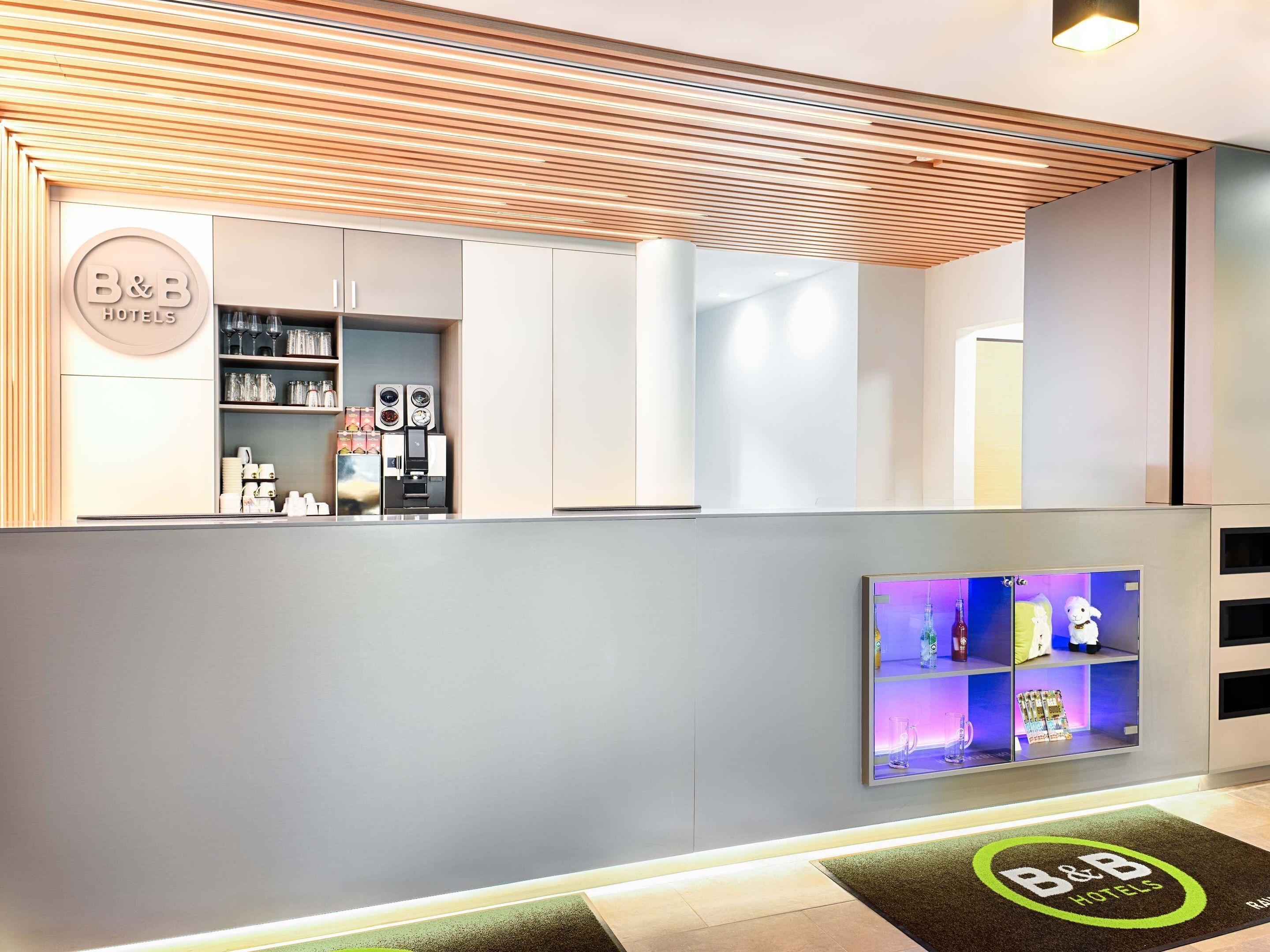 A B&B hotel reception area with a modern design, including a reception desk, a coffee machine, and a display of amenities.
