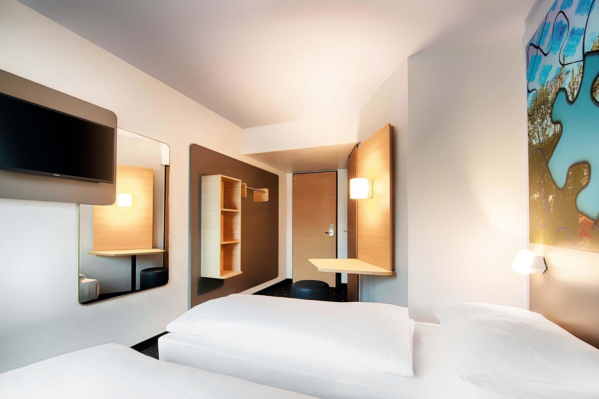 A modern hotel room with two beds, a flat-screen TV, a wooden wardrobe, and a painting on the wall.