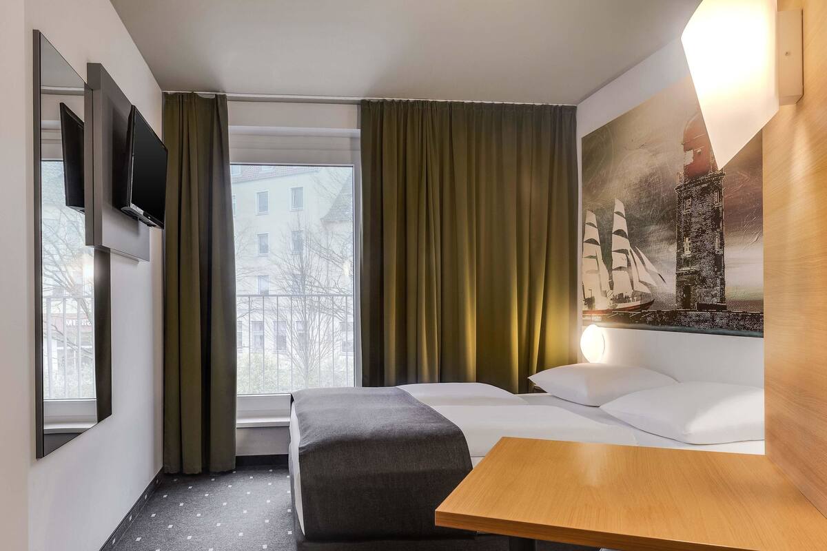 Une chambre d’hôtel avec un lit, une petite table, un canapé, un téléviseur fixé au mur, une fenêtre avec des rideaux et un tableau accroché au mur.