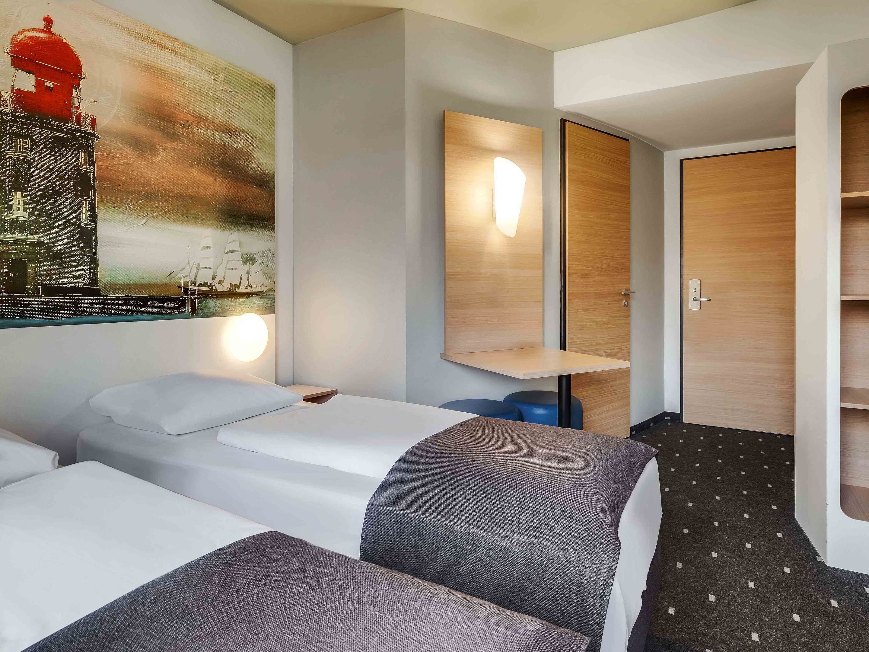 Habitación de hotel con dos camas, un cuadro grande en la pared, una puerta de madera y una mesita.