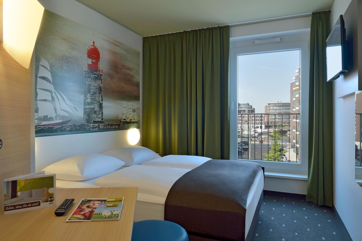 Et hotelværelse med en seng, et skrivebord med en fjernbetjening og blade, et billede af en bygning og et vindue med udsigt over byen.
