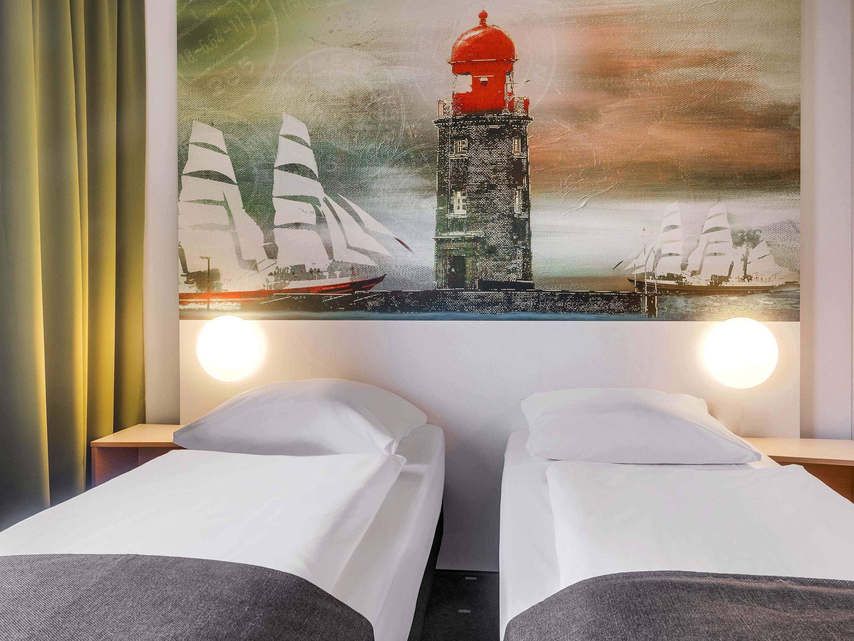 Ein Hotelzimmer mit zwei Einzelbetten, einem großen Gemälde mit einem Leuchtturm und Segelbooten an der Wand sowie zwei Nachttischlampen.