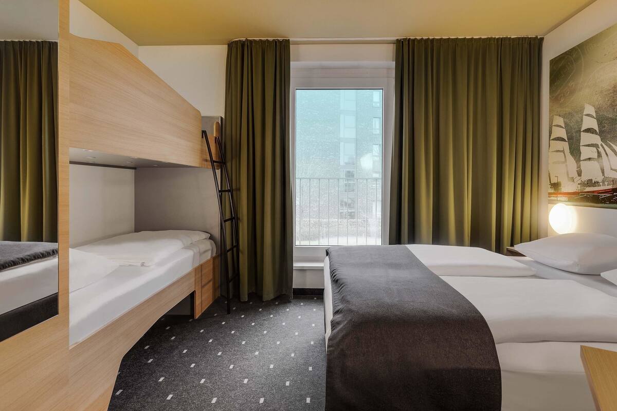 Une chambre d’hôtel avec deux lits, une fenêtre avec des rideaux, un tableau représentant un navire et une vue sur le paysage urbain.