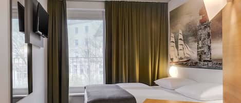 Habitación de hotel con cama, una mesita, un sofá, televisor montado en la pared, una ventana con cortinas y un cuadro en la pared.