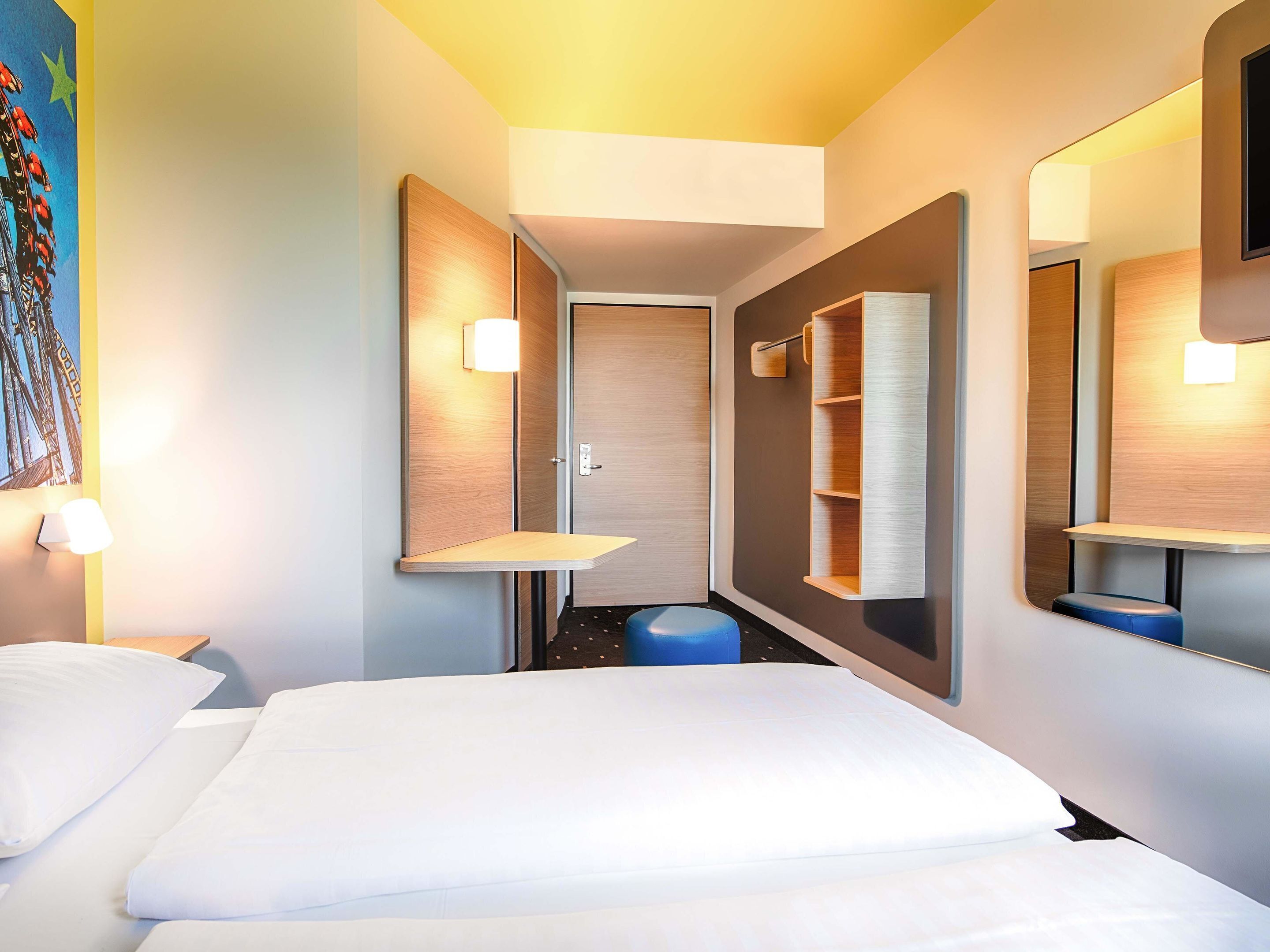 A hotel room with a bed, a desk, a chair, a small stool, a wall-mounted shelf, and a door.