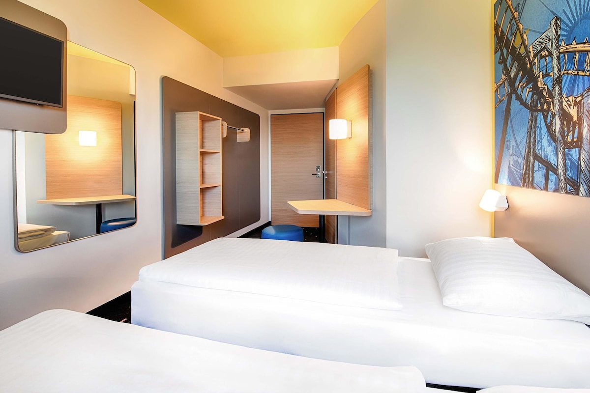 Ein Hotelzimmer mit zwei Betten, einem Badezimmer mit Waschbecken und Spiegel sowie einem Bild an der Wand.