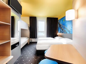 A hotel room with bunk beds, a desk, a chair, a window with curtains, and a wall-mounted lamp.