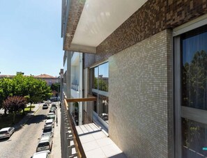 A modern building with a tiled facade, large windows, and a balcony overlooking a street with parked cars and trees.