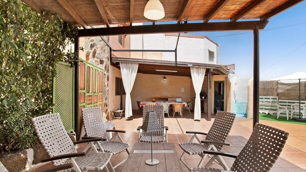 Un área de estar al aire libre con techo, sillones reclinables y una mesa, un muro de piedra y un edificio al fondo.