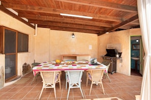 A covered patio with a table set for four, surrounded by wicker chairs, a grill, and a laptop.