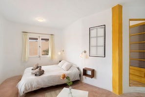 A bedroom with a bed, a window with curtains, a bedside table, a wall-mounted light, and a framed picture.