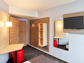 Una habitación de hotel moderna con un armario de madera, una estantería empotrada, un televisor de pantalla plana y un taburete rojo.