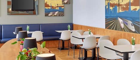 Un restaurante moderno con una larga barra de madera, sillas blancas y una zona de asientos con asientos tapizados en azul. En las paredes, se exhiben pinturas que representan trenes y paisajes urbanos.