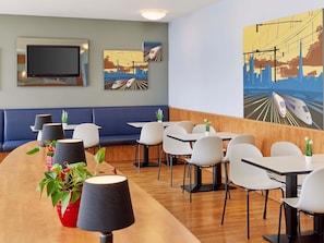 A modern restaurant with a long wooden counter, white chairs, and a blue booth seating area. There are paintings on the walls depicting trains and cityscapes.
