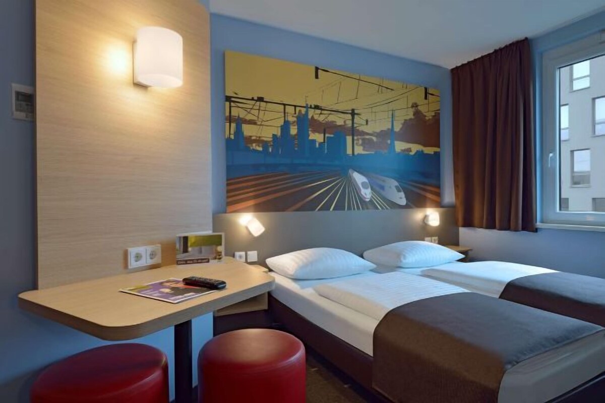 A hotel room with a bed, a desk, two red stools, a painting, and a window with curtains.