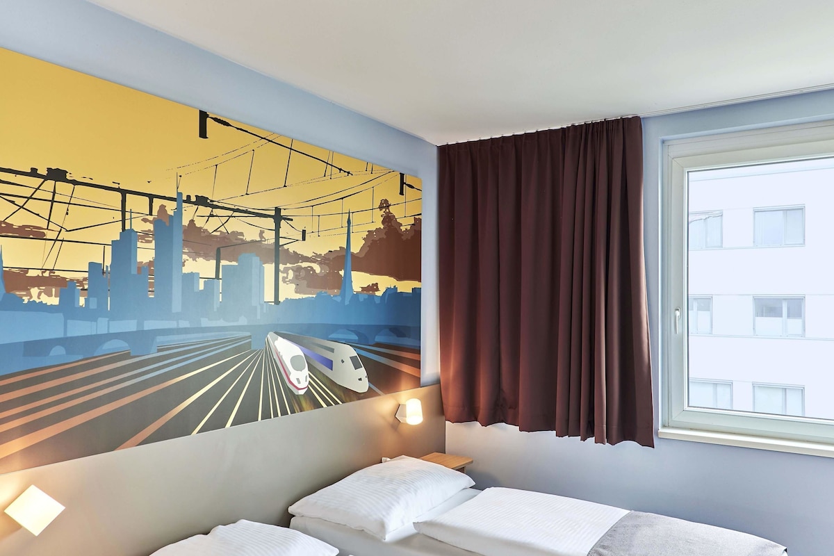 A hotel room with a bed, a window with curtains, a wall mural of a train, and a nightstand with a lamp.