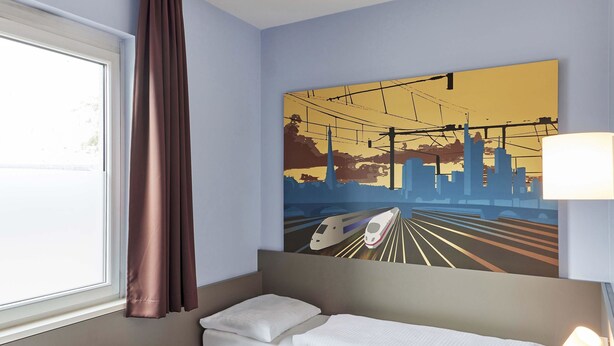 A hotel room with a bed, a window with curtains, a wall-mounted painting of a train, and a bedside lamp. - B&B Hotel Saarbrücken-Hbf (Saarbrücken)