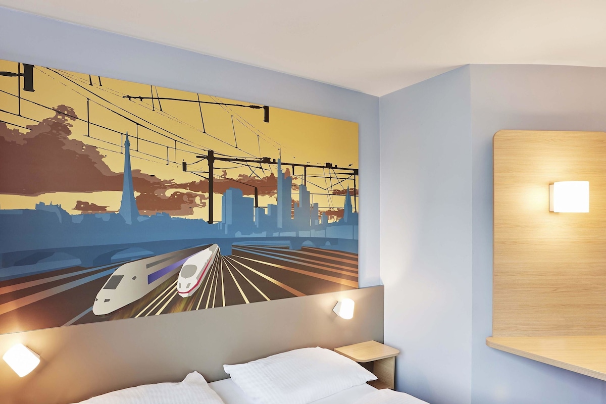 A hotel room with a bed, a nightstand, a wall-mounted light, and a cityscape mural.