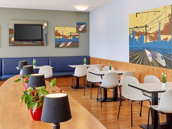 Un restaurant moderne avec un long comptoir en bois, des chaises blanches et un coin salon avec des banquettes bleues. Les murs sont ornés de tableaux représentant des trains et des paysages urbains.
