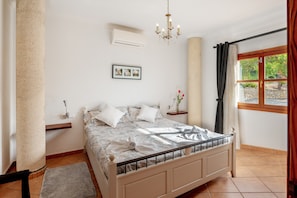 A bedroom with a bed, a chandelier, a window with curtains, a picture on the wall, and a small table with a vase.