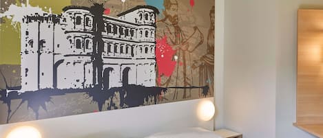 Habitación de hotel con cama, mesita de noche, lámpara de pared y un cuadro abstracto de gran tamaño.