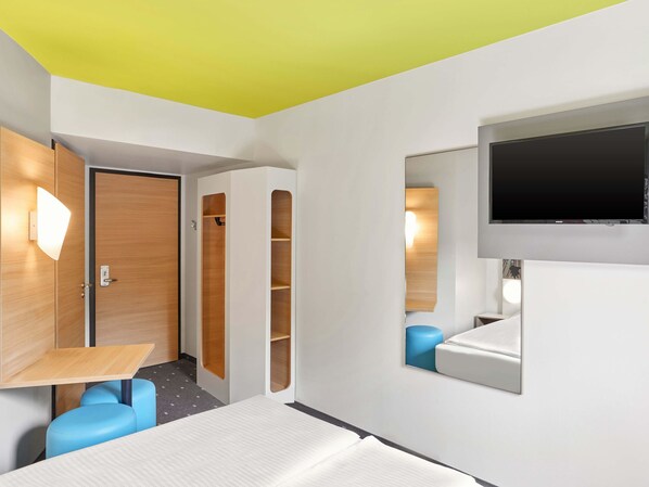 Una habitación de hotel moderna con una cama, un televisor montado en la pared, un armario de madera y una silla azul.