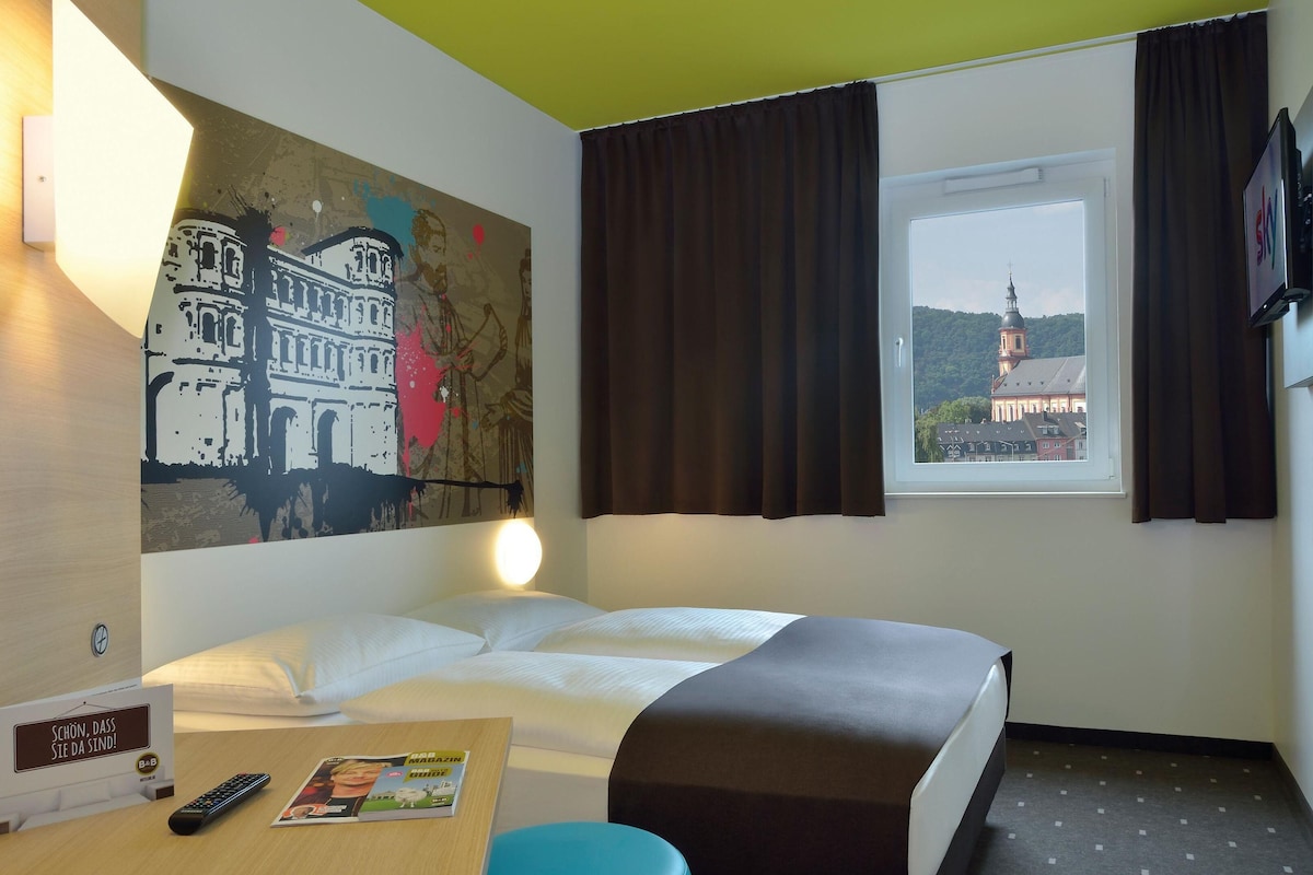 Ein Hotelzimmer mit einem Bett, einem Schreibtisch mit Fernbedienung, einer Zeitschrift und einem Gemälde eines Gebäudes.