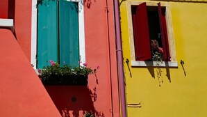 Un edifici amb dues finestres, una amb persianes turquesa i l'altra amb persianes vermelles, i un petit balcó amb flors.