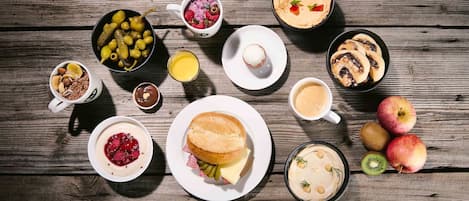 Diversos itens de pequeno-almoço, incluindo panquecas, um sanduíche e frutas frescas como maçãs e kiwi, sobre uma mesa de madeira.
