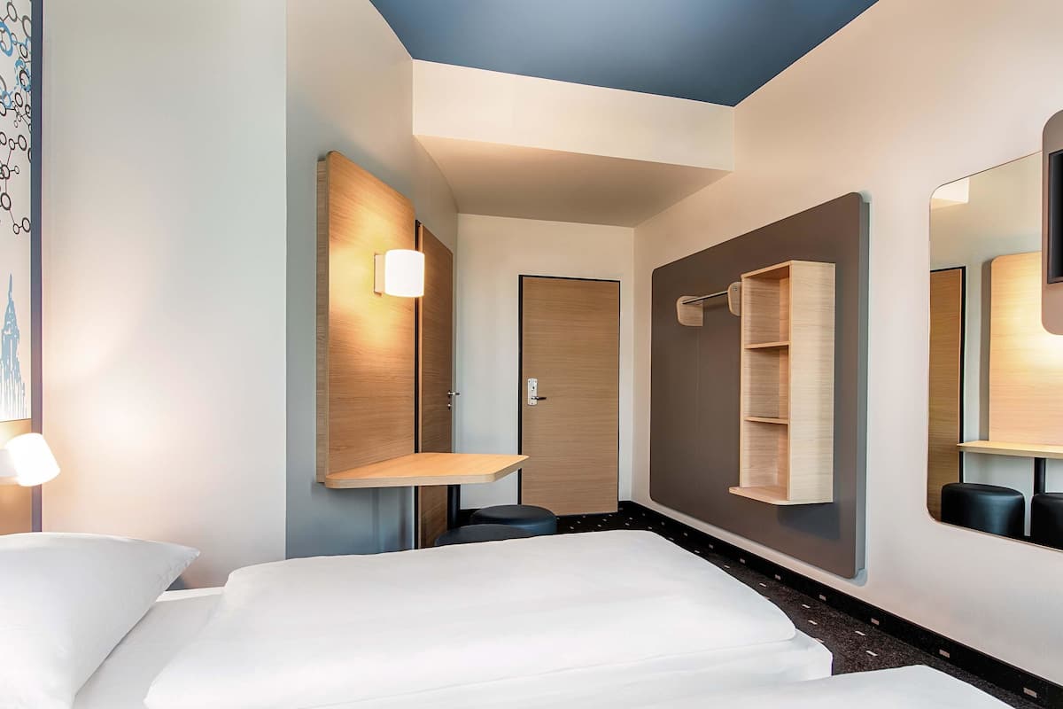 Quarto de hotel com cama, escrivaninha, espelho e porta.