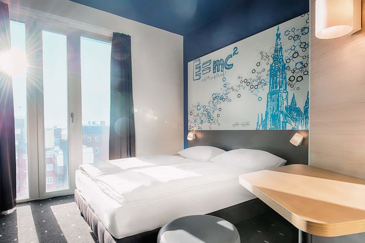 Quarto de hotel com cama, escrivaninha e uma janela grande com cortinas.