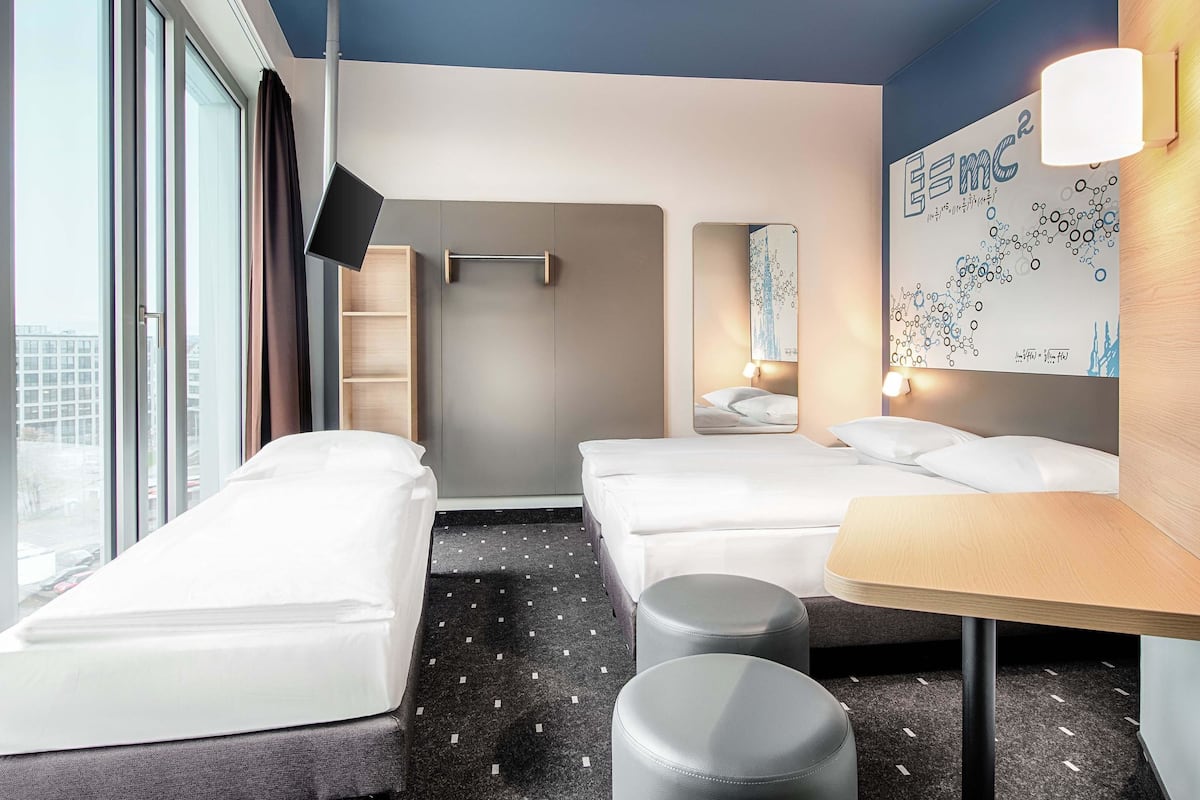 Quarto de hotel moderno com duas camas de solteiro, uma mesinha e uma janela grande.