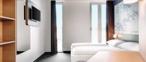 Quarto de hotel com cama, escrivaninha, televisão e janela com cortinas.
