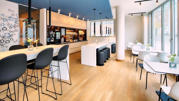A modern bar area with a long counter, black bar stools, and a seating area with white tables and chairs. - B&B Hotel Ulm (Ulm)