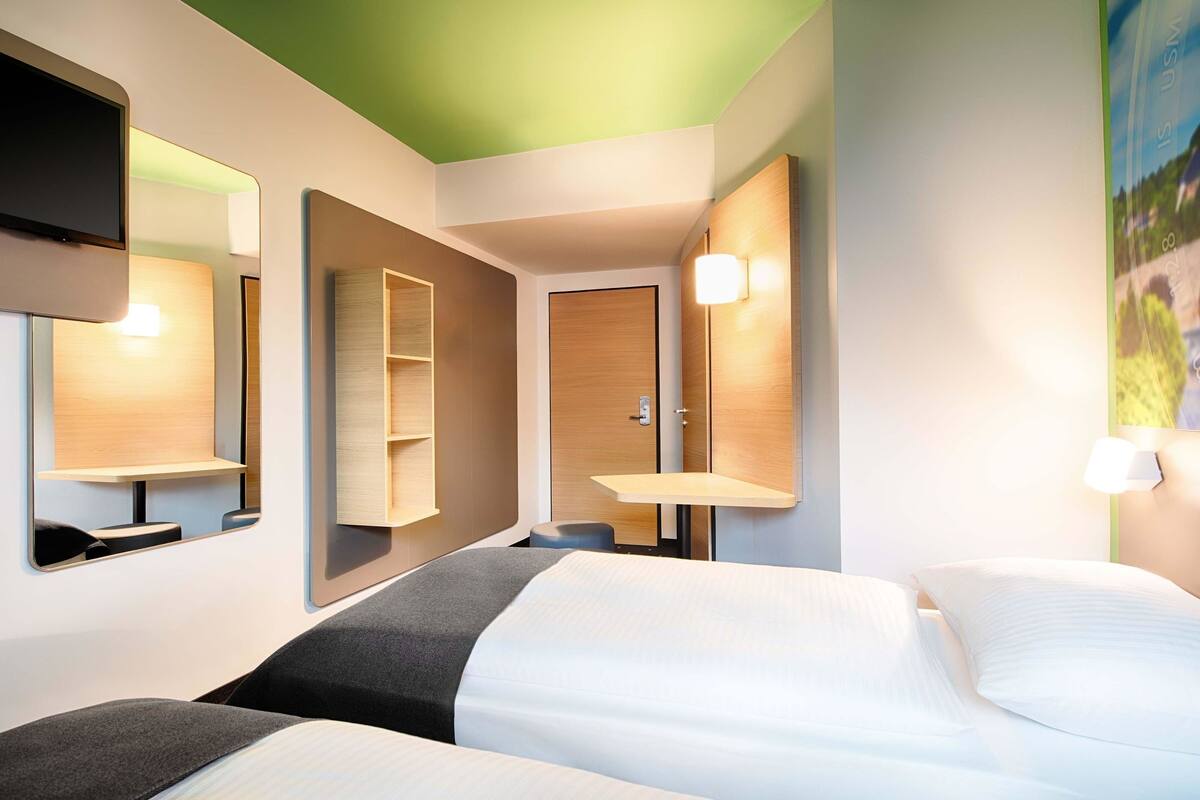 A hotel room with a bed, a bedside table, a mirror, and a wall-mounted shelf.