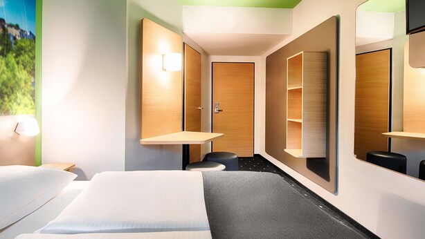 A hotel room with a bed, a desk, a chair, a mirror, and a wardrobe.
