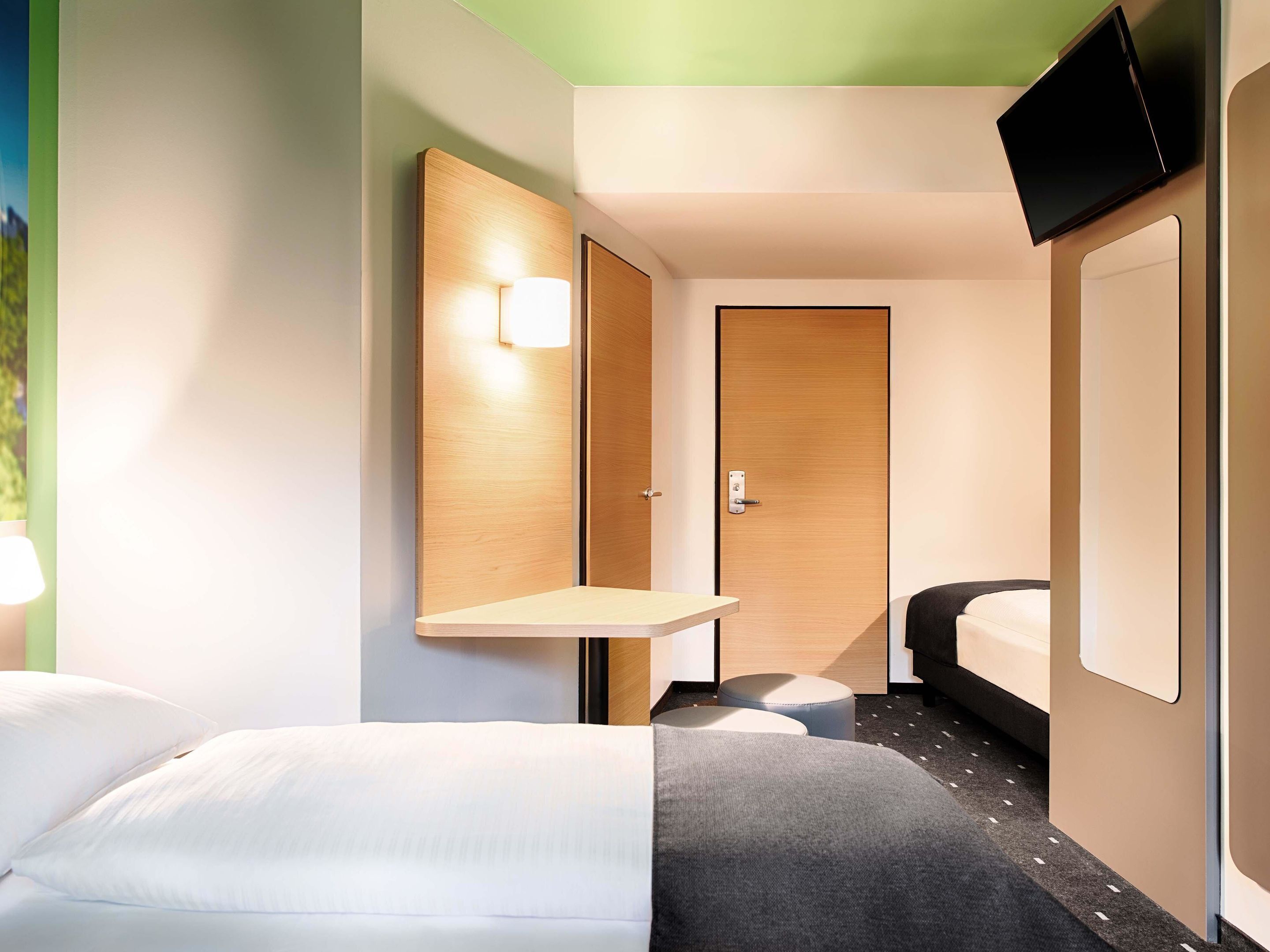 A hotel room with a bed, a bedside table, a TV mounted on the wall, and a door.