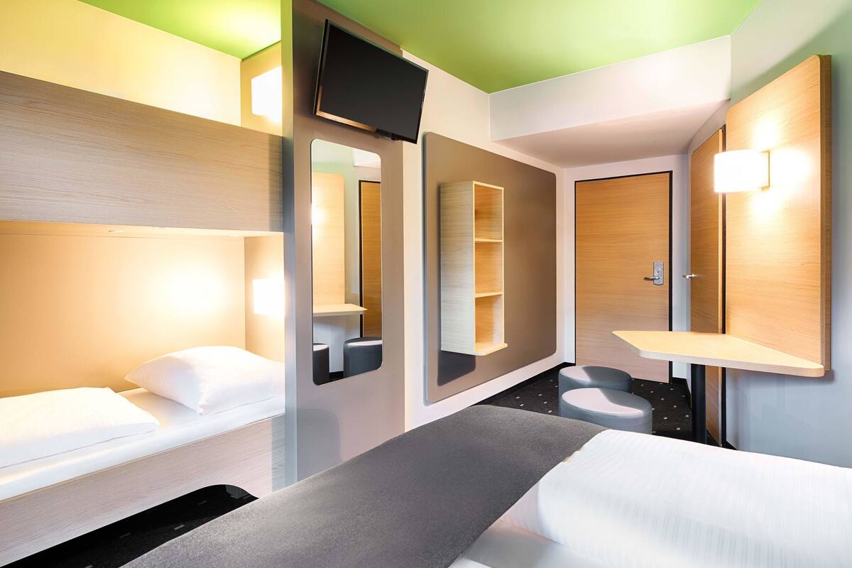 A modern hotel room with a bed, a desk, a mirror, and a television.