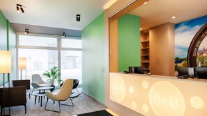A hotel reception area with a desk, chairs, a potted plant, and a view of the outside through large windows.