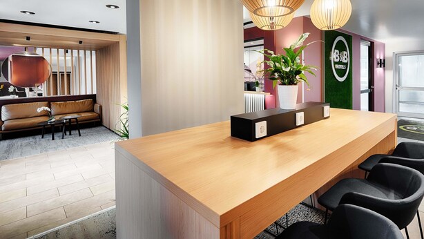 Ein moderner Hotel-Empfangsbereich mit Holztresen, schwarzen Sesseln und einer Zimmerpflanze.