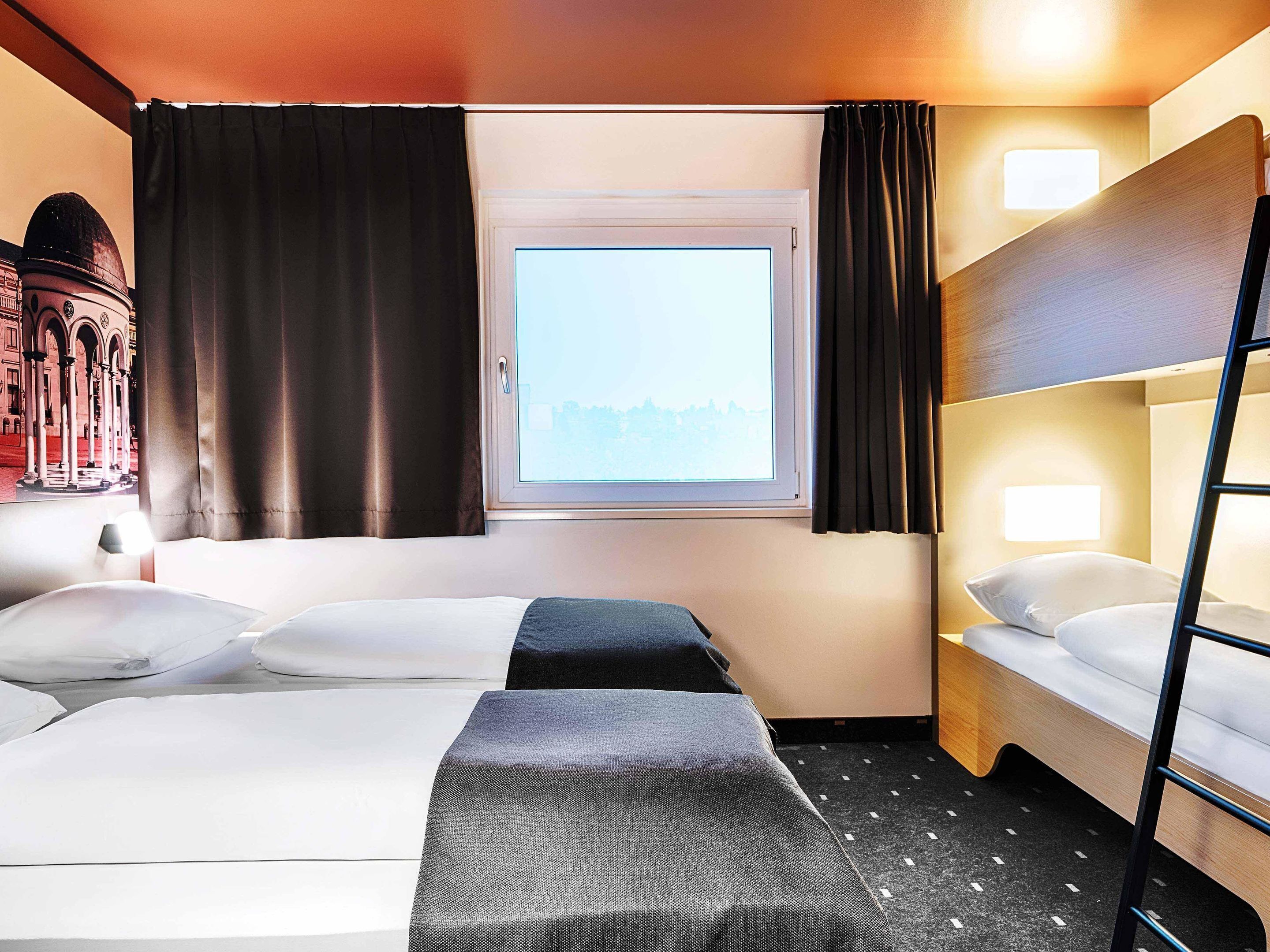 Habitación de hotel con dos camas, ventana con cortinas, lámpara y vista a un edificio.