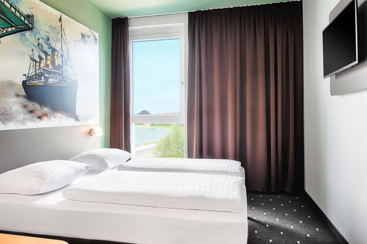 Une chambre d’h ôtel avec un lit, un tableau représentant un navire, une télévision et une vue sur une étendue d’eau, visible par la fenêtre.