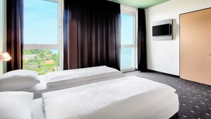 A hotel room with two beds, large windows, a TV, and a view of the city. - B&B Hotel Wilhelmshaven (Wilhelmshaven)