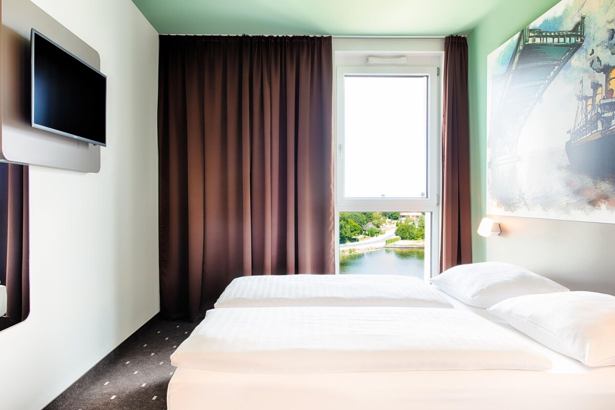A hotel room with a bed, a flat-screen TV mounted on the wall, a window with curtains, and a painting of a bridge.