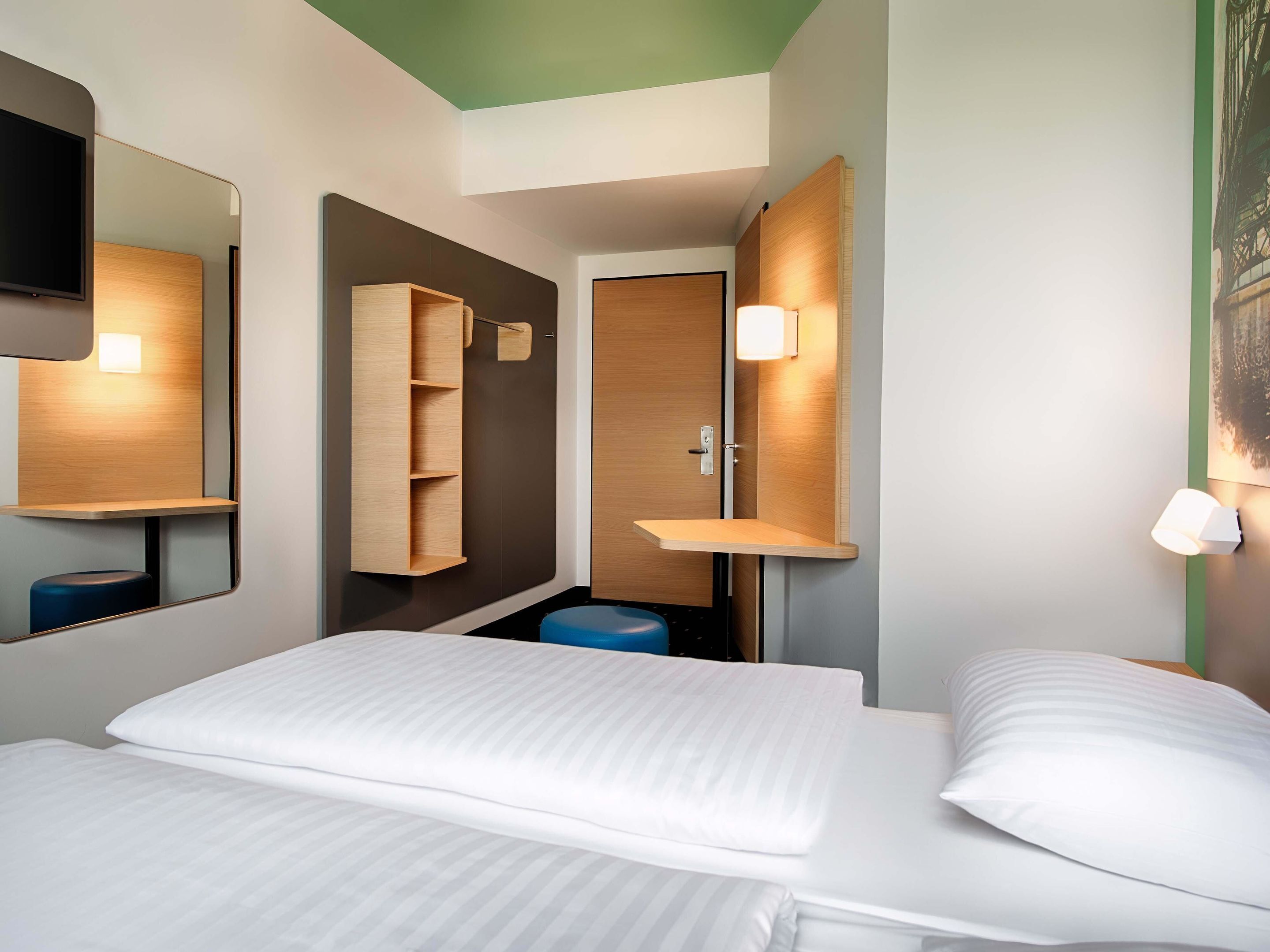 Habitación de hotel con cama, espejo, repisa de madera, lámpara de pared y puerta.