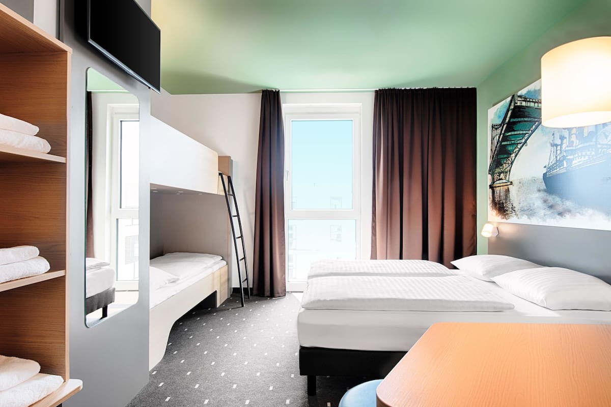 Une chambre d’hôtel avec un lit superposé, un bureau, une chaise, une grande fenêtre avec des rideaux et un tableau au mur.