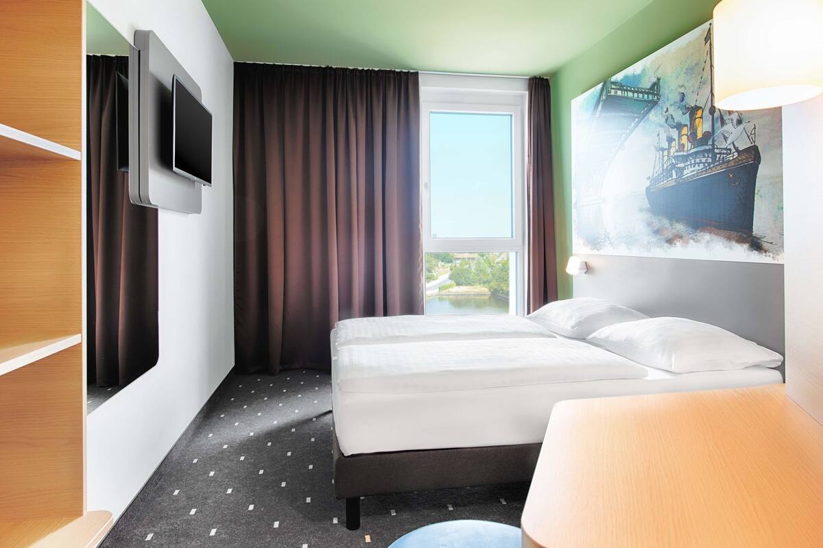 Une chambre d’hôtel avec un lit, un téléviseur fixé au mur, une fenêtre avec des rideaux et un tableau accroché au mur.
