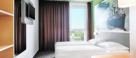 Una habitación de hotel con cama, televisión montada en la pared, ventana con cortinas y un cuadro en la pared.