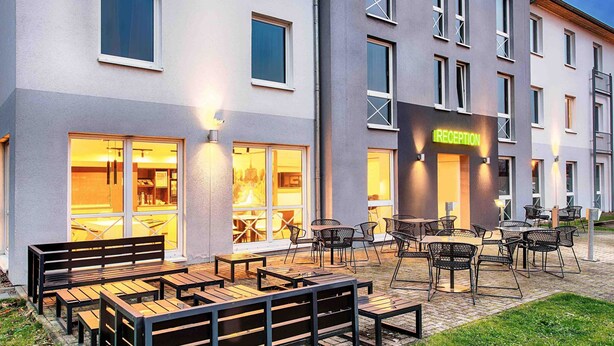 An outdoor seating area with wooden benches and tables, a reception desk, and a building with multiple windows and a 'RECEPTION' sign. - B&B Hotel Kassel-Süd (Lohfelden)